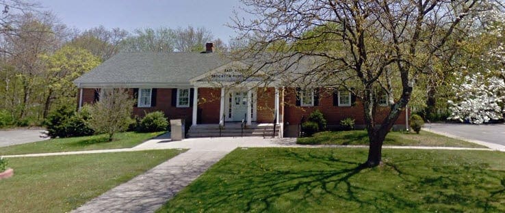 Single-story red-brick building with white columns and a front ramp, surrounded by trees and grass on a sunny day.