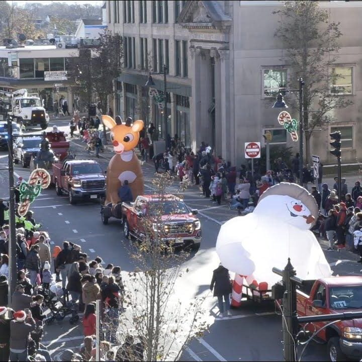 A photo of the Brockton holiday parade. People line the sidewalks while trucks haul parade floats and participants walk alongside.