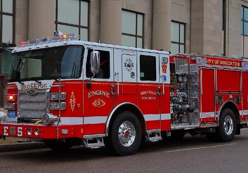A red and white Brockton Fire Department engine truck is parked along a city street in front of a large stone building with tall columns. The truck’s lights are off, and equipment compartments and detailing are visible along its side.