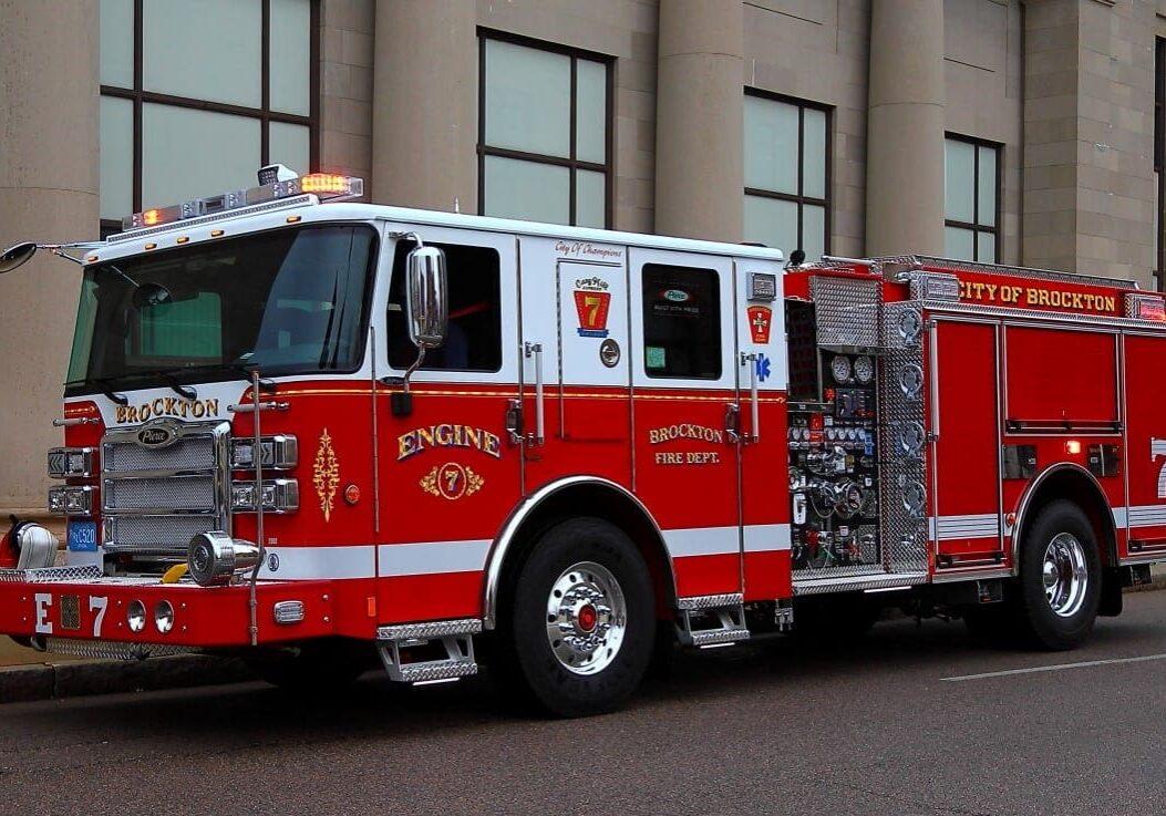 A red and white Brockton Fire Department engine truck is parked along a city street, with its lights, equipment panels, and large front grille clearly visible in front of a stone building.
